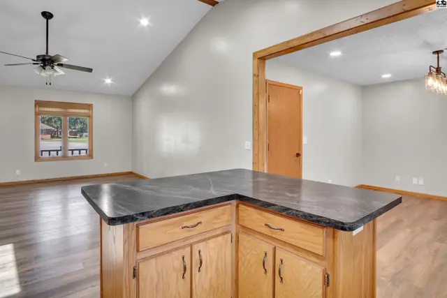 A kitchen island with a dark countertop and wood cabinetry opens to a living area.