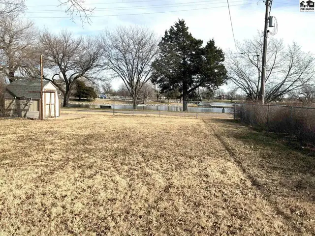 Fenced yard with a storage shed and mature trees borders a body of water.