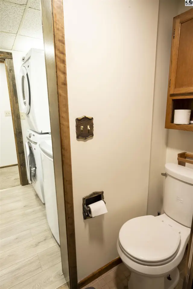 This bathroom features a toilet, wooden cabinetry, and a stacked washer and dryer visible through an open doorway.