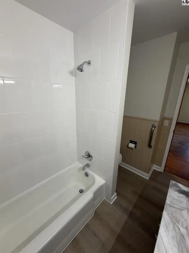 White tiled shower walls surround a bathtub with chrome fixtures and a wood-look floor.