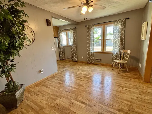 This living area features hardwood floors, a ceiling fan, and large windows with patterned curtains.