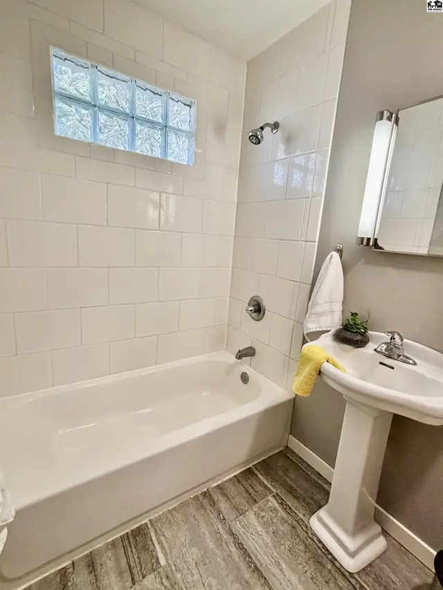 White subway tile surrounds a bathtub, with a pedestal sink and wood-look flooring.