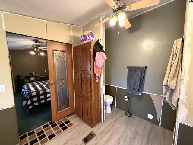 This bathroom features a toilet, wooden cabinet, and ceiling fan, with a doorway to a bedroom.