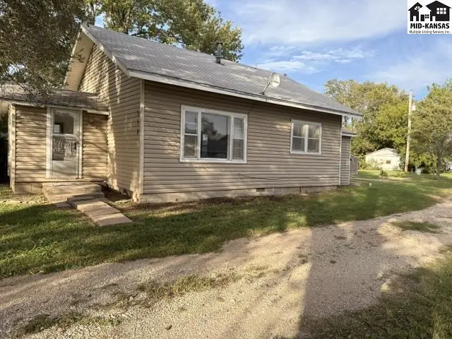 This tan vinyl-sided house features a front porch and mature trees.