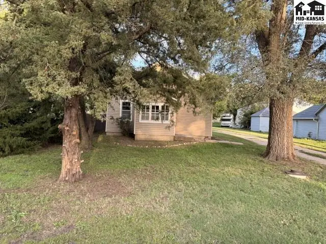 This tan-sided house features a white-trimmed window and mature trees in the yard.