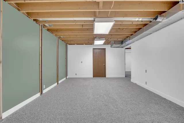 Unfinished basement with carpeted floor, exposed wood ceiling, and drywall.