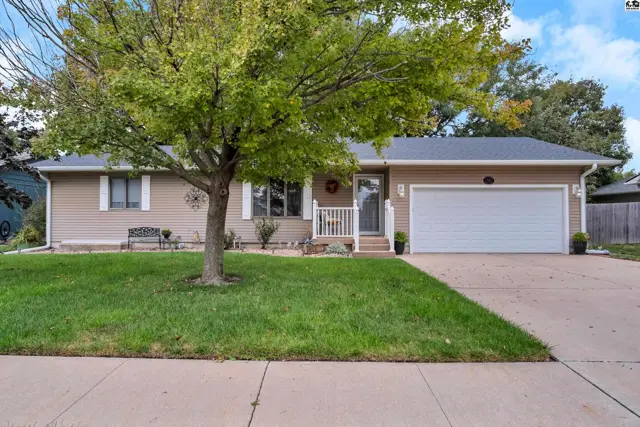This tan-sided home features a white-trimmed front porch, attached garage, and mature tree.
