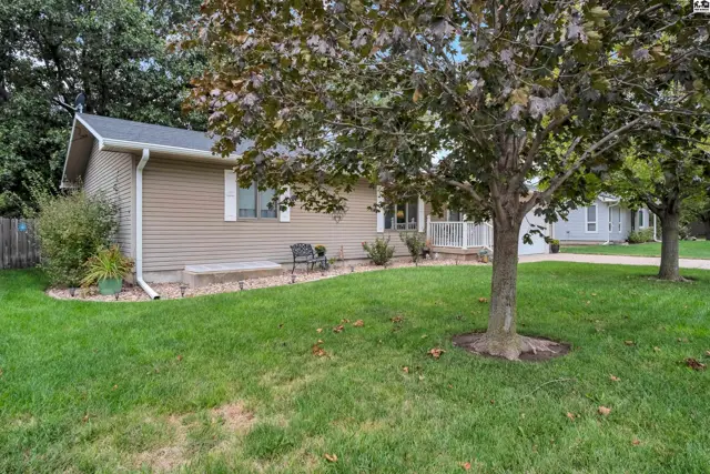 This tan vinyl-sided home features a covered front porch and mature trees.