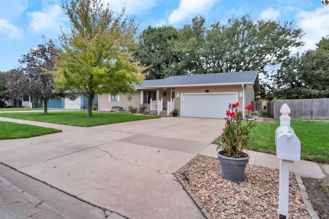 This tan stucco home features a paved driveway, attached garage, and mature trees.