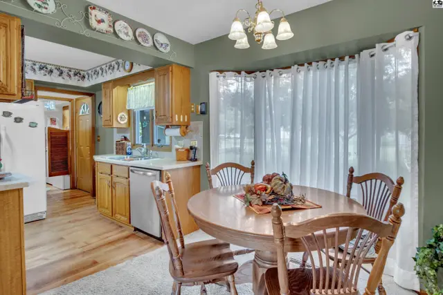 This kitchen features wood cabinetry, a dining table with chairs, and a large window with white curtains.