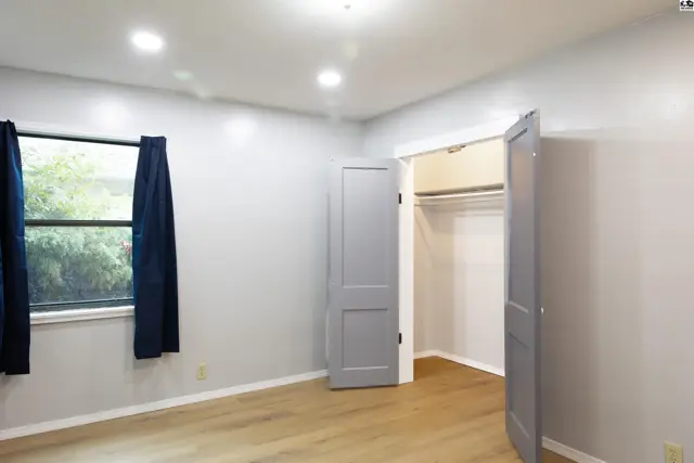 This room features light wood flooring, a large window with dark curtains, and a closet with double doors.