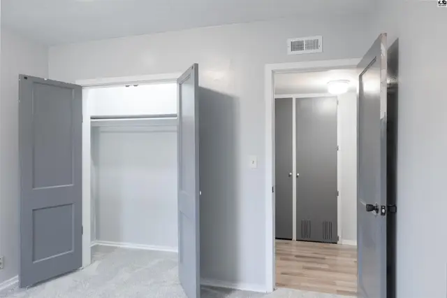 Two open grey doors reveal a closet with a hanging rod and light grey carpet.
