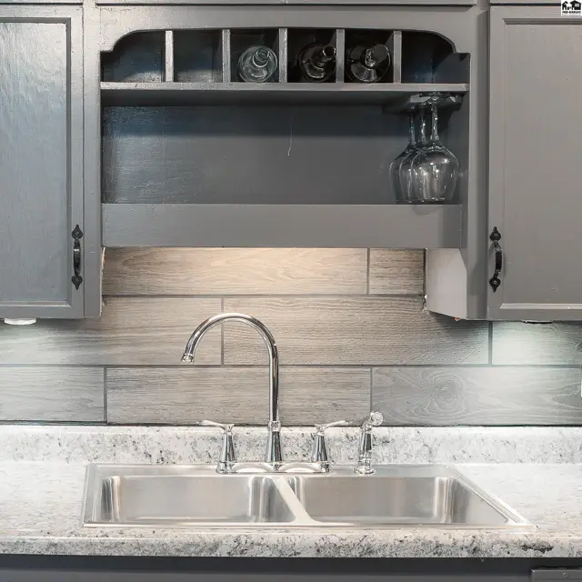 Stainless steel double sink with chrome faucet beneath grey cabinets and wood-look backsplash.