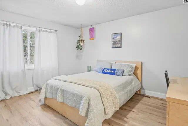 This bedroom features a queen-size bed, light wood flooring, and a window with white curtains.