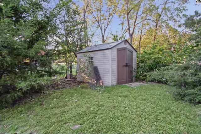 A small, light-colored shed with brown double doors sits in a grassy yard surrounded by trees.