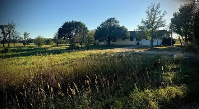 A light-colored house with a garage sits on a large, grassy lot with scattered trees.