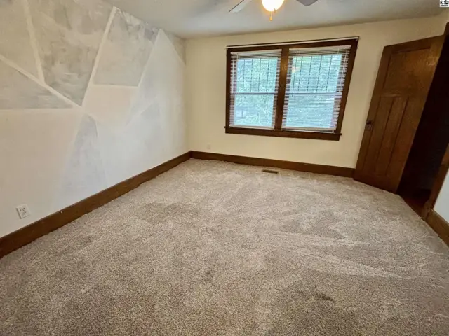 Carpeted room with a ceiling fan, wood-trimmed windows, and a wooden door.