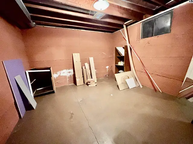 Unfinished basement room with concrete floor, exposed wooden beams, and a small window.