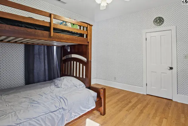 Wooden bunk beds sit on a hardwood floor beneath a patterned wallpapered ceiling.