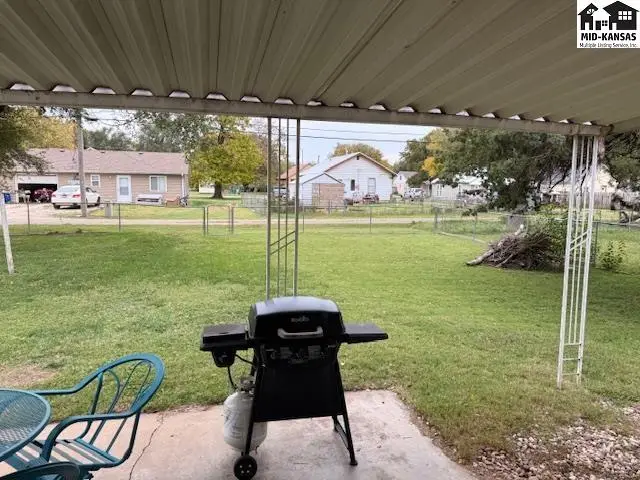Covered patio with a grill overlooks a fenced yard with grass and trees.
